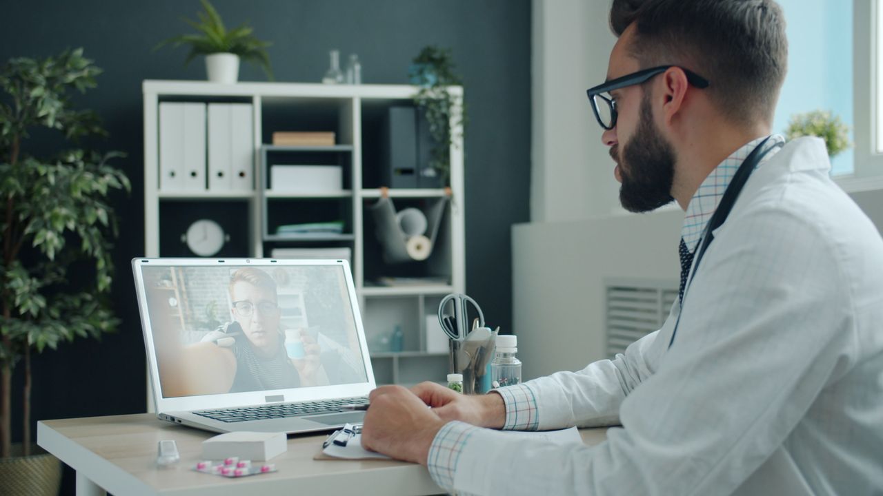 Services Doctor in white coat video calling a patient on a laptop in a modern office setting.